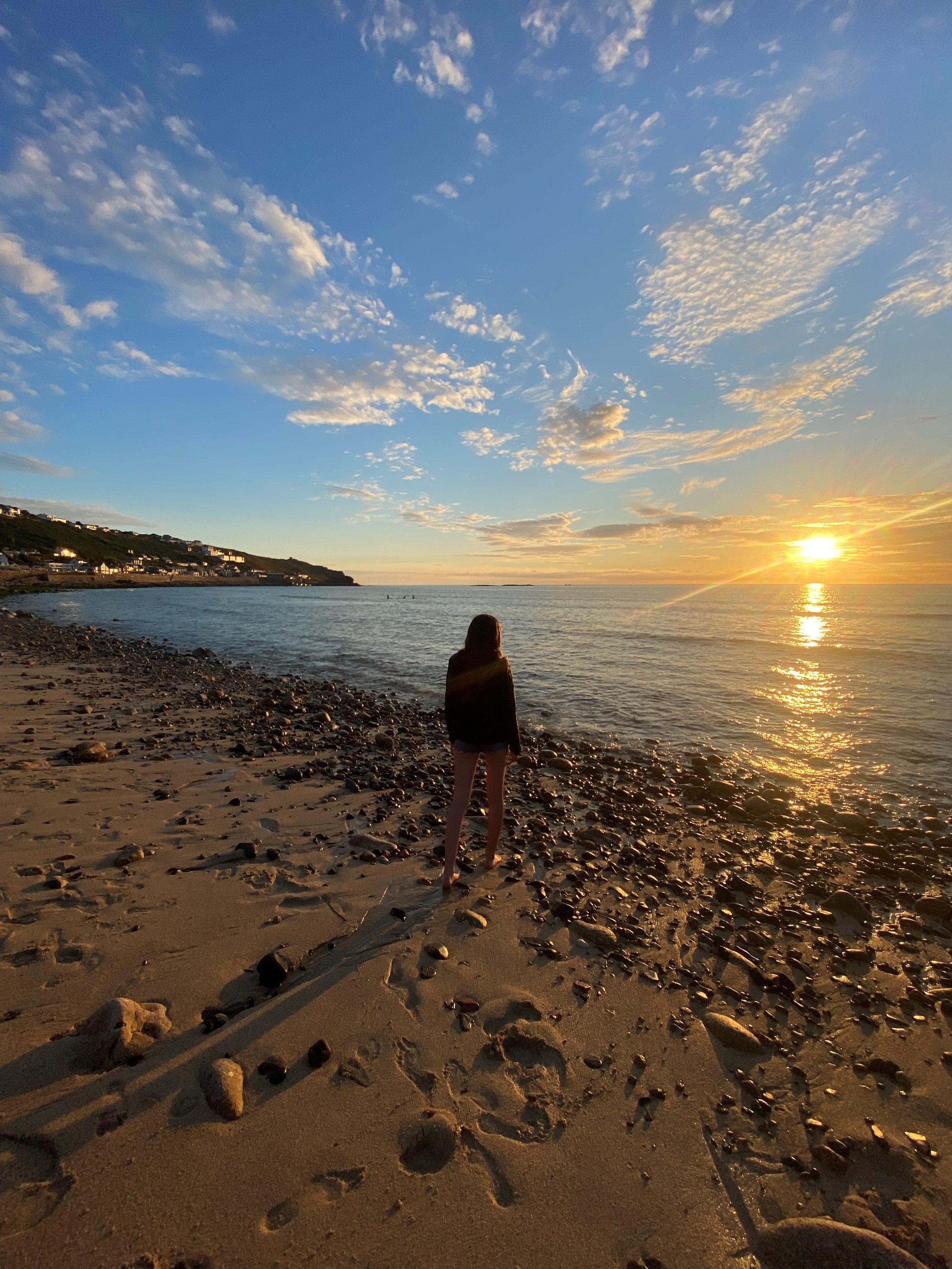 the sun dips into the sea at Sennen beach as a lone figure stands and watches the sunset